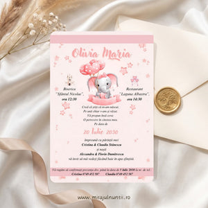 Invitație de botez cod Magnific B.13-20013 cu fond roz pal, elefănțel gri cu fundiță roz care ține baloane-inimioare roz cu buline, steluțe roz și benzi roz, numele Olivia Maria, așezată pe satin alb lângă un plic crem cu sigiliu auriu din ceară