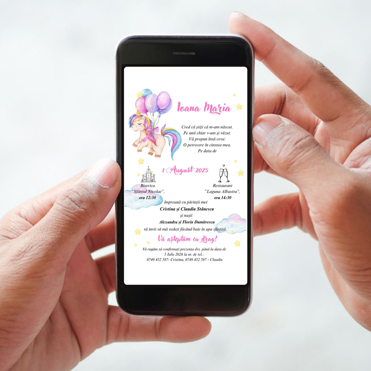 Invitație digitală de botez cod Magnific B.15-20015 afișată pe ecranul unui telefon ținut în mâini: unicorn în acuarelă cu coamă curcubeu și baloane roz și lila, numele Ioana Maria scris caligrafic cu roz, nori pastel și steluțe galbene