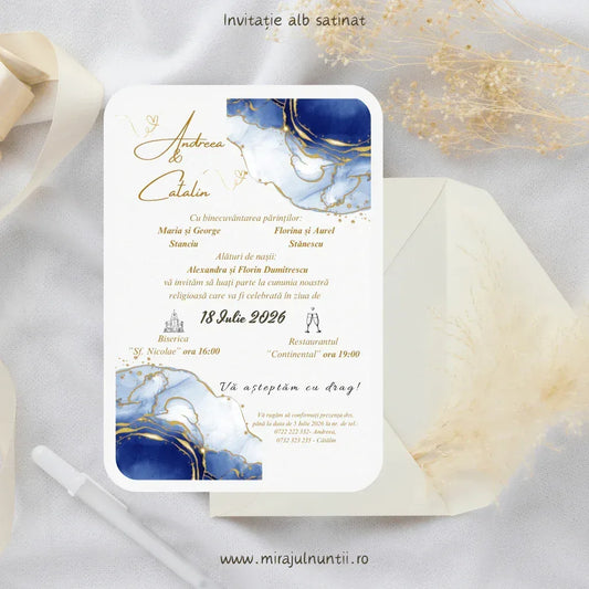 Invitație de nuntă cod Magnific C.3 pe carton alb satinat, cu acuarelă bleumarin și vene aurii în colțuri și numele mirilor scrise caligrafic cu auriu, lângă un plic crem și ierburi pampas.