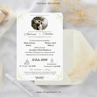 Invitație de nuntă cod Magnific C.2 pe carton alb satinat, cu fotografia mirilor în partea de sus, frunze aurii fine în colțuri și nume scrise caligrafic, lângă un plic alb și ierburi pampas.