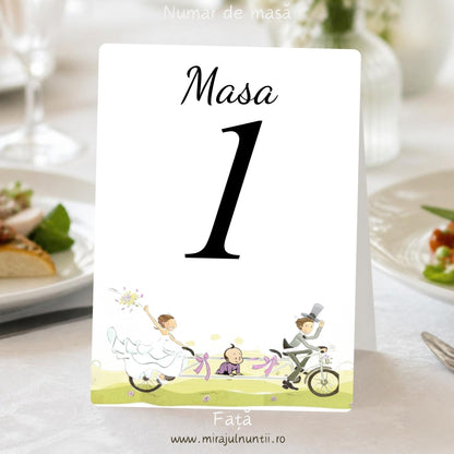 Număr de masă alb, față: „Masa 1” negru, iar jos mirii pe bicicleta tandem cu funde lila, bebelușul de-a bușilea și iarbă verde-fistic, cod Miraj 20371