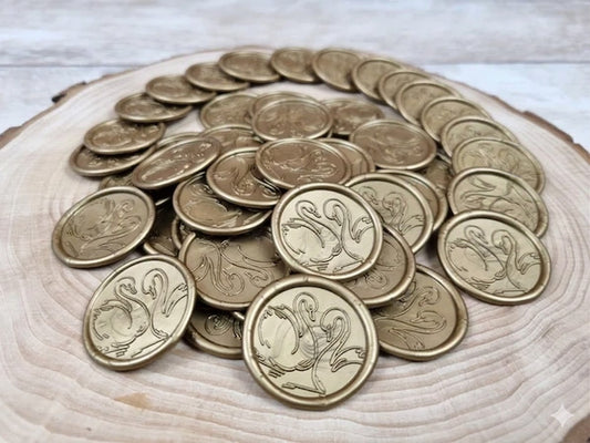 Set 50 de sigilii din ceară naturală de culoare auriu metalizat, cu design reliefat reprezentând două lebede, așezate pe un suport decorativ din lemn.